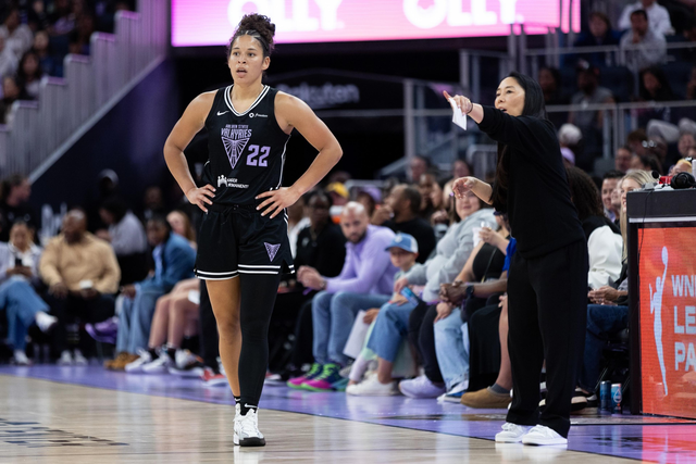 A basketball player stands on the court in a black uniform with "22" on it, while a coach gestures and speaks from the sidelines. Spectators watch in the background.